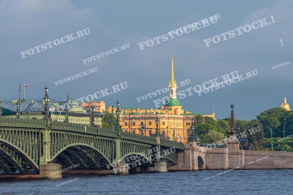 На фотографии изображён вид на Троицкий мост и Инженерный замок в Санкт-Петербурге. Свет освещает архитектурные детали, выделяя золотистый шпиль на фоне ясного неба. Это фото может использоваться в туристических брошюрах, образовательных материалах о Санкт-Петербурге, статьях о российской архитектуре или как изображение для постов в социальных сетях, посвященных путешествиям.